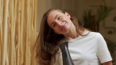 Beyaz tişörtlü genç bir kadın banyodaki aynaya bakarken saç kurutma makinesi kullanıyor ve gülümsüyor.