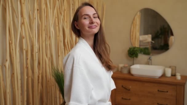 Smiling young woman with perfect smooth skin looking at the camera, an attractive girl in a bathrobe standing in the bathroom — Video Stock