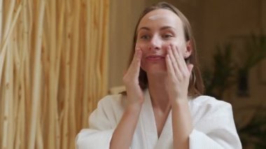 Bornozlu, gülümseyen güzel bir kadın yüzüne krem sürüyor ve banyo aynasına bakıyor.