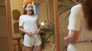 İnce bir kadın aynanın önünde duruyor ve kilo verdikten sonra belini ölçüyor.