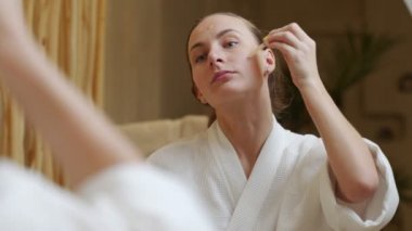 Güzel genç bir kadın aynaya bakar ve banyoda yüzüne serum uygular.