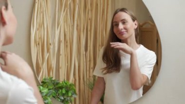 Genç bir kadın banyoda dikilirken yüzüne dokunuyor ve gülümsüyor.