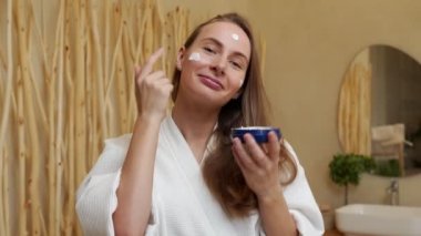 Bornozlu, gülümseyen güzel bir kadın yüzüne krem sürüyor. Bir kadın banyo aynasına bakıyor. Sabah sağlıklı cilt bakımı