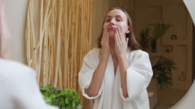 Bornozlu güzel bir kadın banyoda dikiliyor ve yüzünü aynanın önünde losyonla temizliyor, makyajını temizliyor.