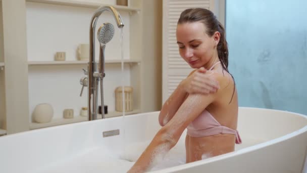 Young woman applying an exfoliating scrub on her shoulder after a shower while sitting in the bathroom — стоковое видео