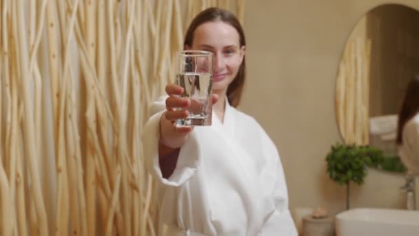 Hermosa joven en un albornoz sostiene un vaso de agua y sonríe — Vídeo de stock