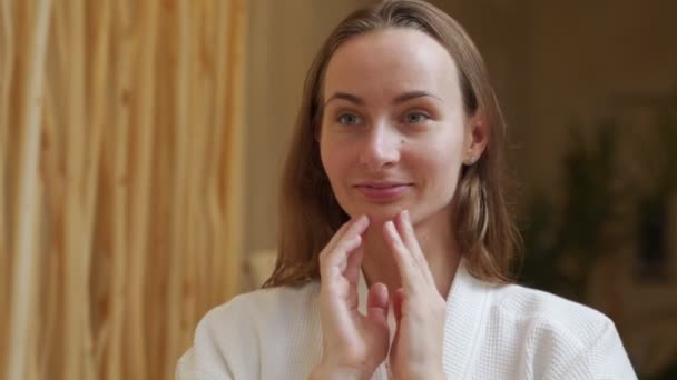Smiling beautiful woman in a bathrobe puts a skin care cream on her face and looks in the bathroom mirror — Wideo stockowe