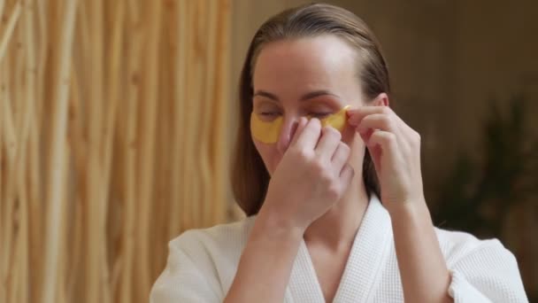 Young woman wearing bathrobe applying cosmetic eye patches while standing at the bathroom mirror — Stock Video