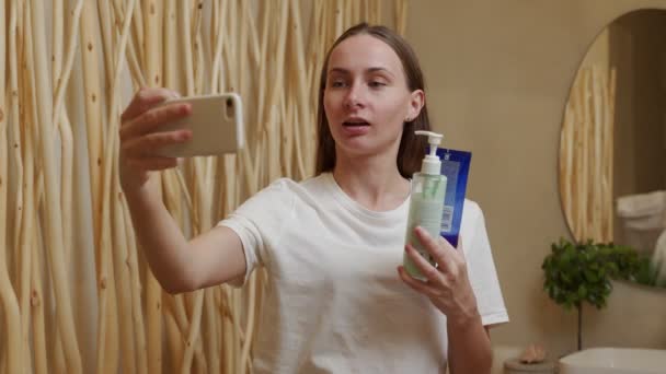 Beauty blog about cosmetics. A happy woman looks at a smartphone webcam and shows her creams in the bathroom — Vídeo de Stock