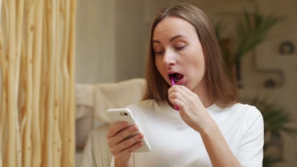 Mulher morena usando smartphone enquanto escova os dentes no banheiro — Vídeo de Stock