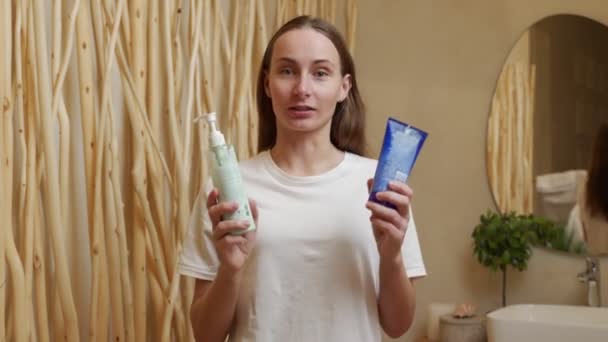 Blog de belleza sobre cosméticos. Una mujer feliz mira a la cámara y muestra sus cremas en el baño — Vídeo de stock