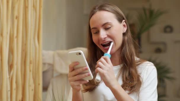 Woman brushing teeth and reading message on phone from bathroom. Girl with smartphone using toothbrush, checking social networks. — Vídeo de Stock