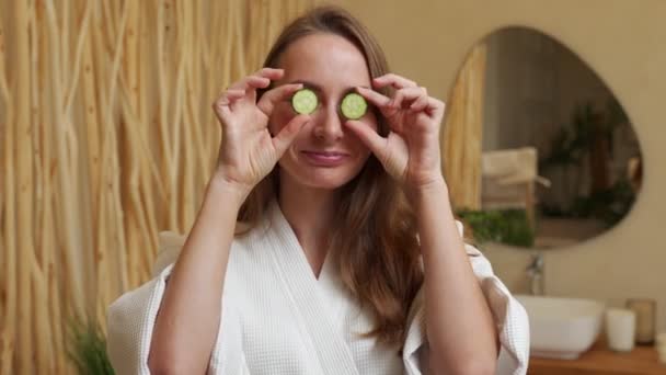 Attractive young woman in a bathrobe holding cucumber slices over her eyes while standing in the bathroom — Stockvideo