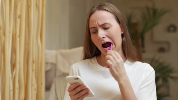 Mujer cepillarse los dientes y leer el mensaje en el teléfono desde el baño. Chica con teléfono inteligente usando cepillo de dientes, comprobando las redes sociales. — Vídeo de stock