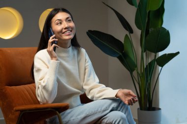 Gülümseyen genç bir kadın evde bir sandalyede otururken akıllı bir telefonla konuşur.