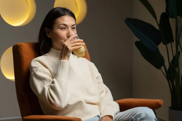 Asiática mujer bebe jugo de naranja de un vaso mientras se sienta en el sofá y relajarse — Foto de Stock