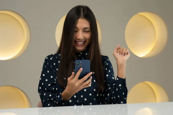 An Asian woman sits at her desk in the office, holds a smartphone, receives a message from the bank and feels incredibly happy. The woman makes a gesture look very excited, winning the lottery — Foto de Stock