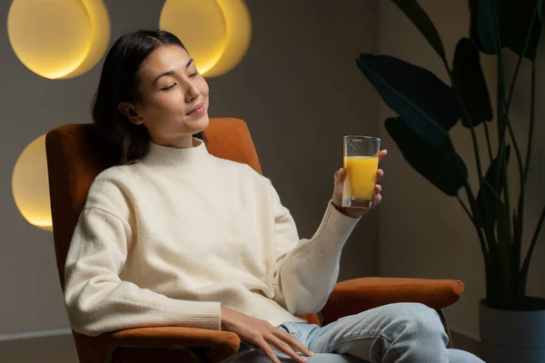 Asiática mujer bebe jugo de naranja de un vaso mientras se sienta en el sofá y relajarse — Foto de Stock