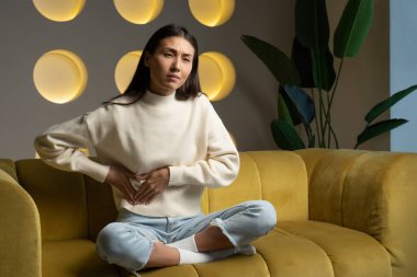 Young Asian woman in a sweatshirt, whose hands touch her stomach, suffers from abdominal pain while sitting on the couch at home