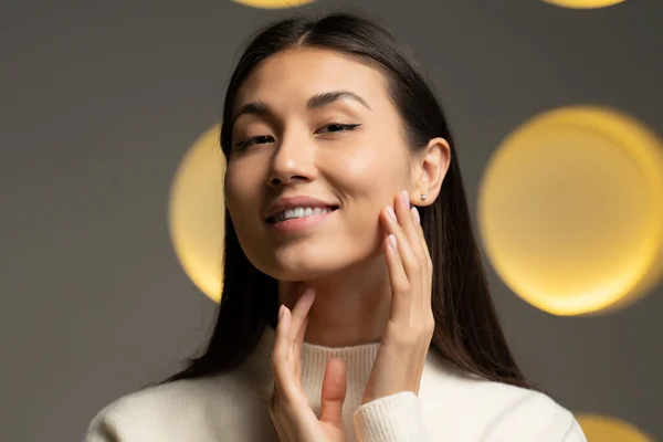 Portrait of a beautiful young Asian woman touching her perfect skin and looking into the camera. Close-up of a womans face massaging her face — Stock Photo, Image