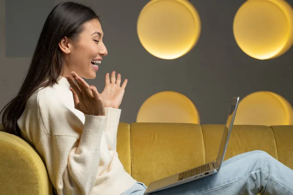 Emocional joven mujer asiática con un ordenador portátil celebra la victoria en un sofá amarillo en casa. Una hermosa joven usa una computadora portátil y sonríe mientras está sentada en el sofá de su casa. — Foto de Stock