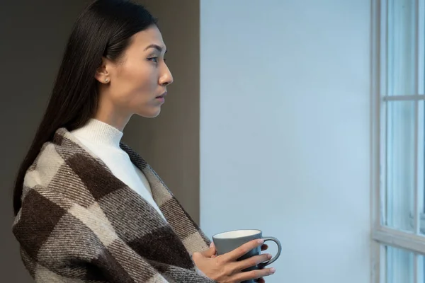 Mujer asiática triste, cubierta con una manta, bebe café o té y mira a la calle desde grandes ventanas modernas — Foto de Stock