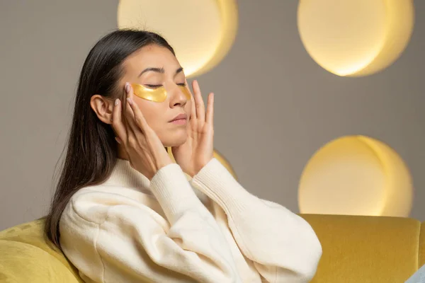 Young Asian woman with clear radiant skin with moisturizing golden patches under her eyes is resting on the sofa in the living room — Stock fotografie