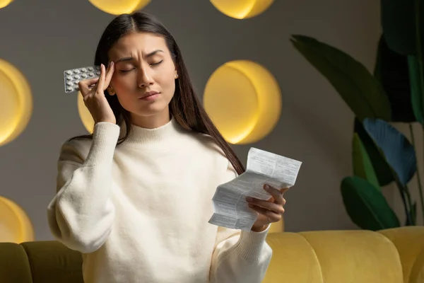 Una mujer asiática toma medicamentos para el dolor de cabeza. Una joven sostiene un paquete de pastillas en su mano y lee instrucciones médicas.. — Foto de Stock