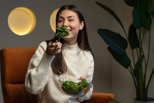 Cheerful young Asian woman who eats fresh salad and smiles sitting on a chair in the living room — стоковое фото