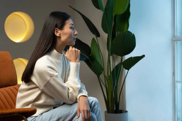 Una joven asiática está sentada dentro de la casa y mirando por la ventana mientras está sentada en una silla. La mujer está decepcionada, triste y molesta. — Foto de Stock