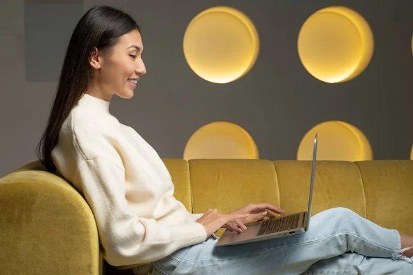 Feliz mujer asiática está sentada en el sofá amarillo de su casa usando un ordenador portátil que navega por la red — Foto de Stock