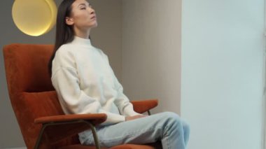 Asian woman in a white sweater sitting comfortably in an armchair, throws her hands behind her head