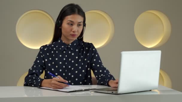 A young Asian woman using a headset looks at a laptop screen, listens and studies online courses. — Vídeo de Stock