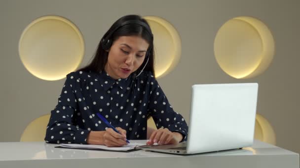 A young Asian woman using a headset looks at a laptop screen, listens and studies online courses. — Vídeo de Stock