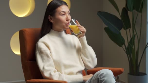 Asian woman drinks orange juice from a glass while sitting on the couch and relaxing — ストック動画