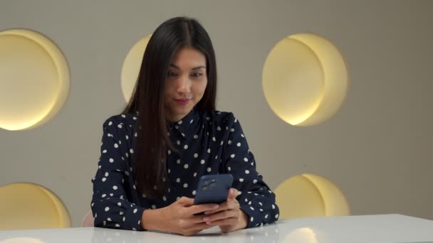 Una mujer asiática se sienta en su escritorio en la oficina, sostiene un teléfono inteligente, recibe un mensaje del banco y se siente increíblemente feliz. La mujer hace un gesto muy emocionado, ganar la lotería — Vídeo de stock