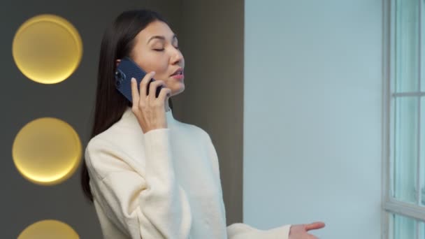 Young Asian woman is talking on a mobile phone and smiling, standing at the window, a freelance woman is calling on the phone, the concept of communication. — Vídeo de Stock