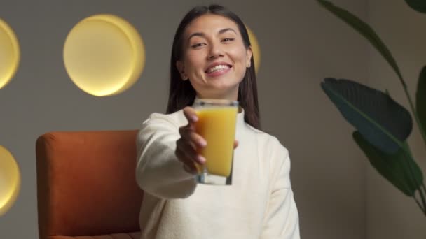 Beautiful Asian woman holds a glass of orange juice in her hand and looks at the camera — Vídeo de Stock
