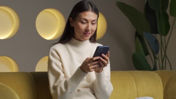 Mujer asiática sonriendo usando el teléfono móvil, mujer sorprendida haciendo un gesto ganador en el teléfono inteligente — Vídeos de Stock