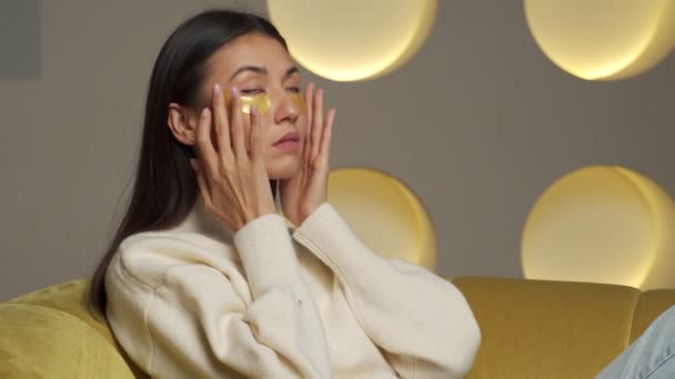 Young Asian woman with clear radiant skin with moisturizing golden patches under her eyes is resting on the sofa in the living room — Wideo stockowe