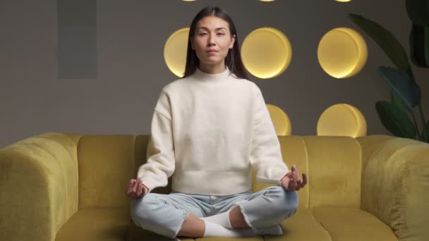 Time for yoga classes. Attractive young asian woman trains and sits in a yoga lotus position on the couch — Wideo stockowe