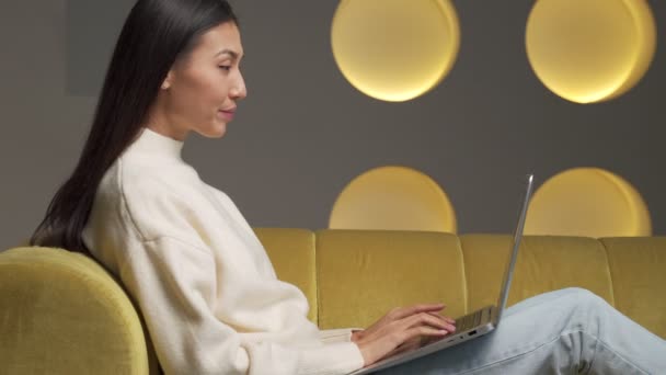 Emotional young Asian woman with a laptop celebrates victory on a yellow sofa at home. A beautiful young woman uses a laptop and smiles while sitting on the couch at home. — Video