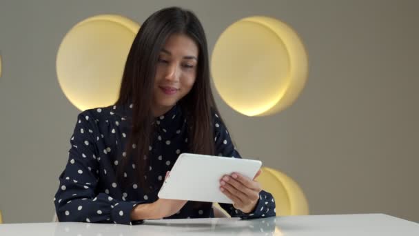 Stylish Asian woman sitting on a table with a tablet in her hands, using the Internet in a modern office with an interior. — ストック動画