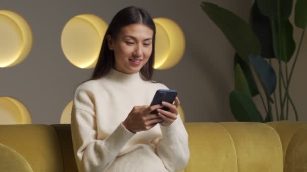 An excited Asian woman waving her fists got good news on her smartphone while sitting on the couch — Vídeos de Stock