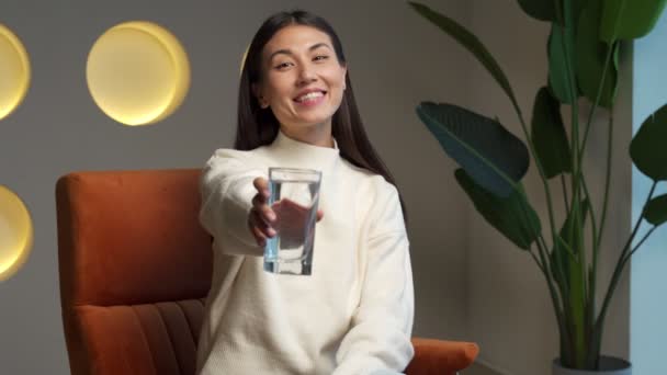 A happy beautiful young Asian woman holding a clear glass of clean water in her hand and looking at the camera. Portrait of a happy smiling young woman with a glass of fresh water — Vídeos de Stock