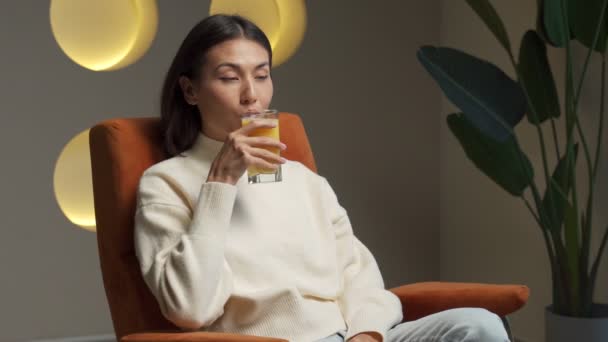 Asian woman drinks orange juice from a glass while sitting on the couch and relaxing — Wideo stockowe
