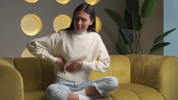 Young Asian woman in a sweatshirt, whose hands touch her stomach, suffers from abdominal pain while sitting on the couch at home — Vídeo de Stock