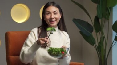 Cheerful young Asian woman who eats fresh salad and smiles sitting on a chair in the living room