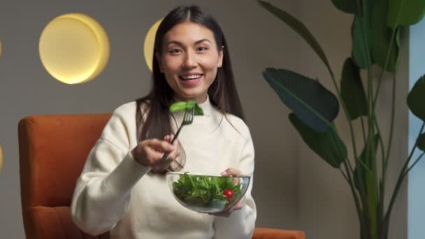 Cheerful young Asian woman who eats fresh salad and smiles sitting on a chair in the living room — Vídeos de Stock