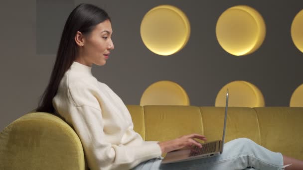 Happy Asian woman is sitting on the yellow sofa of her home using a laptop surfing the net — Wideo stockowe